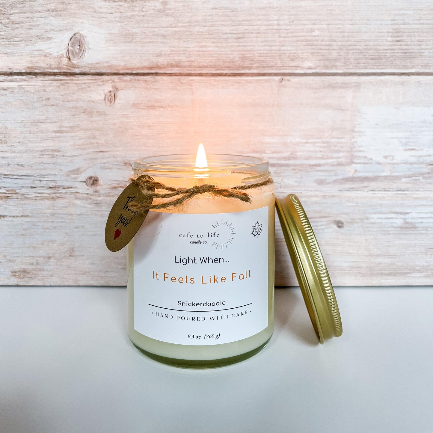Glass candle with flame. Gold top on side. Label says "Light When... It Feels Like Fall"